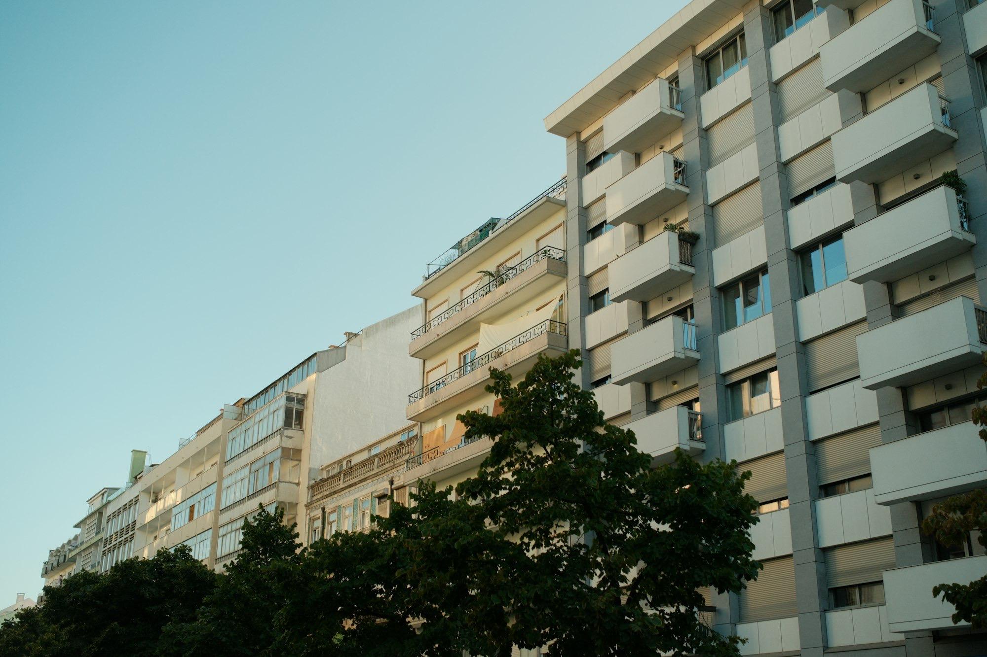 A row of light-coloured apartment buildings with balconies above a green tree canopy in warm sunlight.