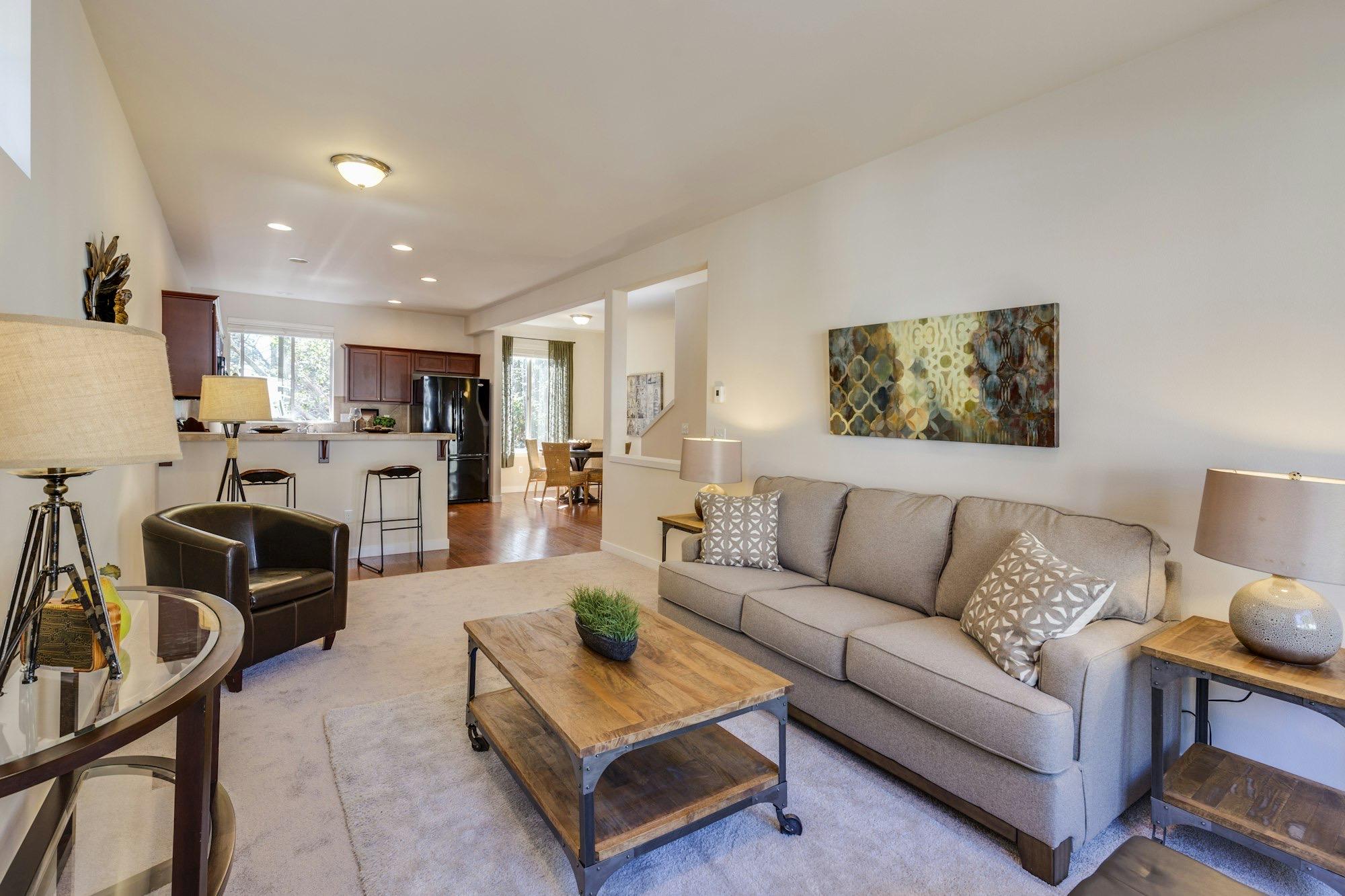 A bright living room with a beige sofa and wooden coffee table, opening onto a kitchen and dining area.