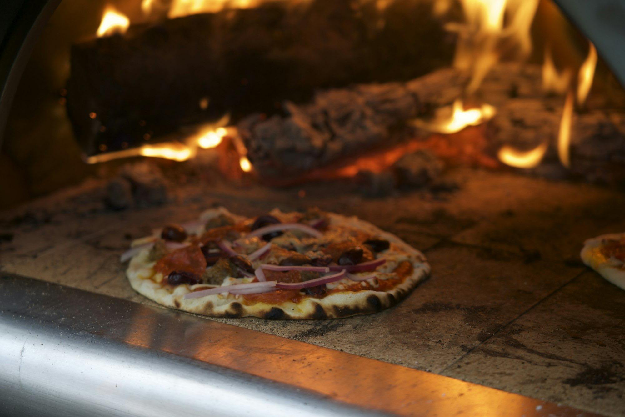A pizza baking on the stone floor of a wood-fired oven, with flames burning behind it.