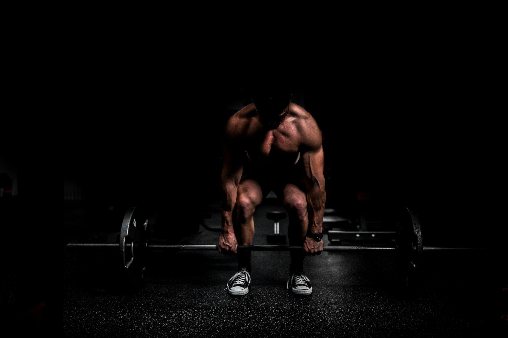 An athlete bent over a loaded barbell in a dark gym, lit from above.